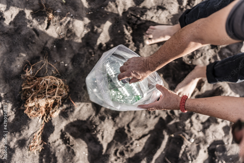 Fototapeta premium Hands of adult man picking up plastics on the beach, collection of polluting waste on the coast, on beaches.