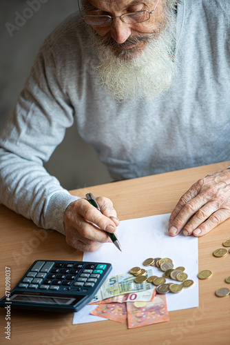 Old bearded senior man with calculator and bills counting euro money and writing notes on white sheet of paper.