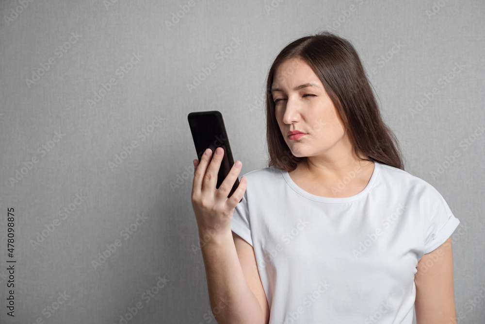 Dark haired young woman with poor eyesight dressed in white t-shirt looks closely at phone screen on gray studio background closeup Dark haired young woman with poor eyesight dressed in white t-shirt looks closely at phone screen on gray studio background closeup