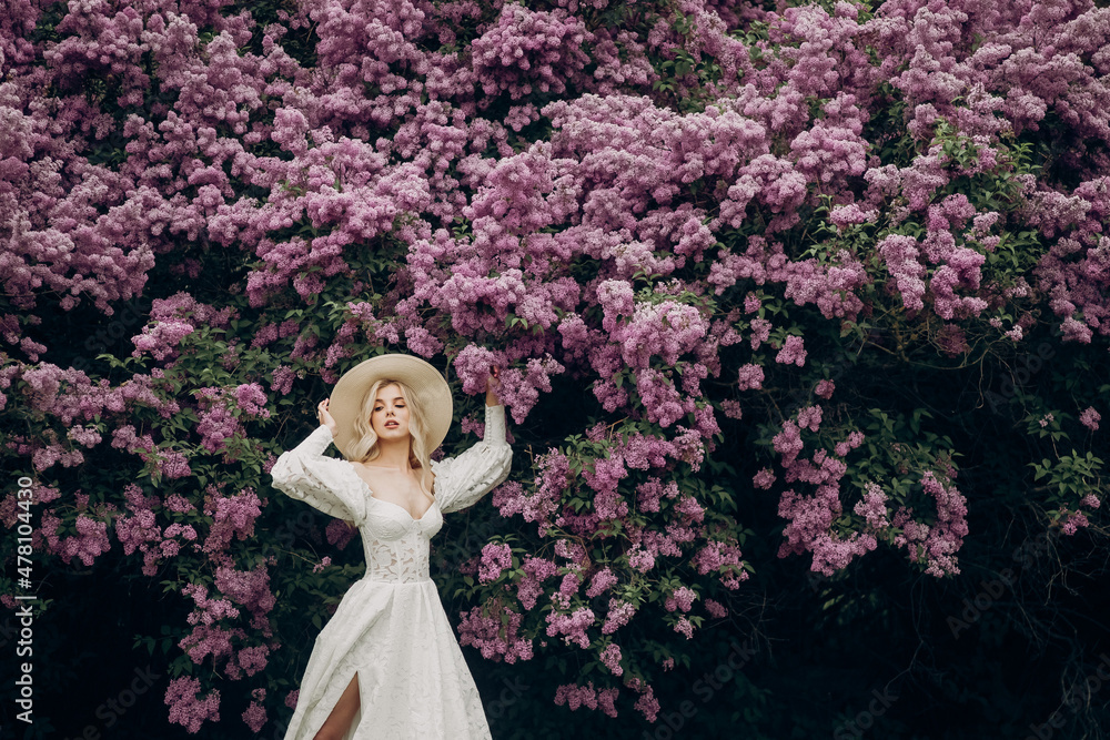 Young blonde girl in a straw hat and in a white wedding dress with an open top near the lilac bushes