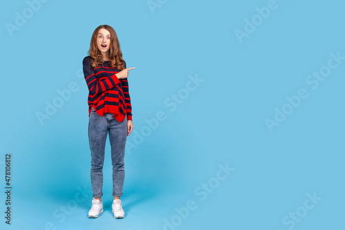 Wow, look, advertise here! Full length portrait of woman showing copy space with shocked expression, showing blank wall for crazy commercial idea. Indoor studio shot isolated on blue background.