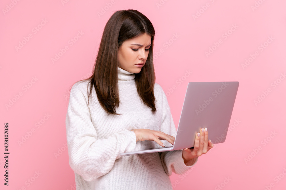 Side view portrait of pensive girl works online, looks at laptop display with thoughtful expression, wearing white casual style sweater. Indoor studio shot isolated on pink background. Side view portrait of pensive girl works online, looks at laptop display with thoughtful expression, wearing white casual style sweater. Indoor studio shot isolated on pink background.