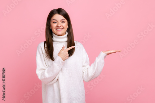 Happy woman presenting copy space on her palm, showing empty place for commercial text or goods, wearing white casual style sweater. Indoor studio shot isolated on pink background.