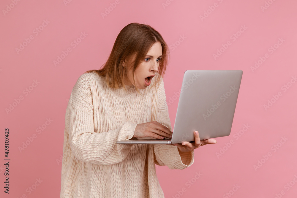 Portrait of amazed shocked young adult blond woman working on laptop compute, looking at display with open mouth, wearing white sweater. Indoor studio shot isolated on pink background. Portrait of amazed shocked young adult blond woman working on laptop compute, looking at display with open mouth, wearing white sweater. Indoor studio shot isolated on pink background.