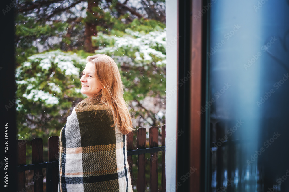 Young happy girl blonde woman on the balcony of her house enjoying the snow and forest view wrapping herself in a checkered plaid Young happy girl blonde woman on the balcony of her house enjoying the snow and forest view wrapping herself in a checkered plaid