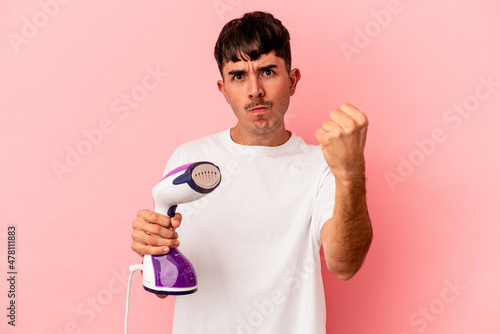 Young mixed race man holding an iron isolated on pink background showing fist to camera, aggressive facial expression.