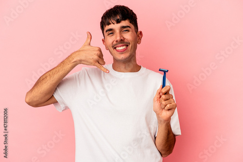 Young mixed race man holding razor blade isolated on pink background showing a mobile phone call gesture with fingers.