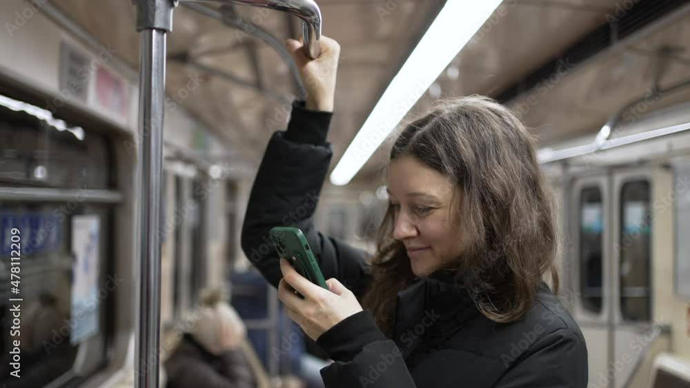 Brunette-haired woman wearing warm puffy jacket scrolls through social media using smartphone and holding on to handrail in subway wagon closeup Brunette-haired woman wearing warm puffy jacket scrolls through social media using smartphone and holding on to handrail in subway wagon closeup
