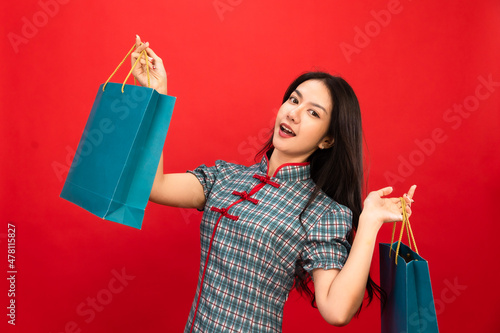 Asian woman in modern cheongsam costume holding shopping bags isolated on red studio background. Chinese new year in shopping concept style.