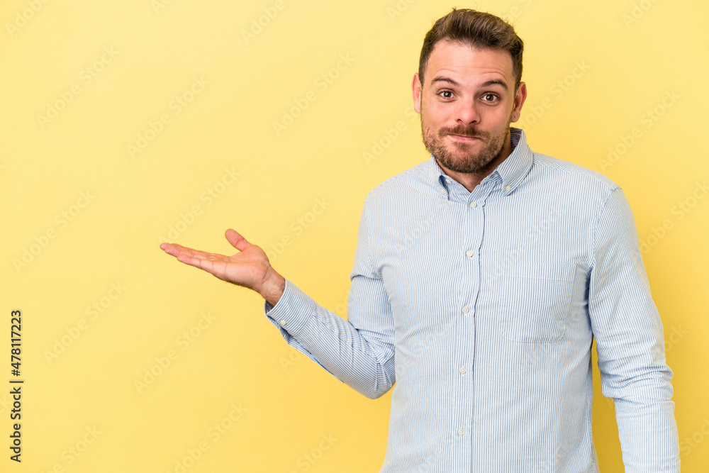 Young caucasian man isolated on yellow background showing a copy space on a palm and holding another hand on waist. Young caucasian man isolated on yellow background showing a copy space on a palm and holding another hand on waist.