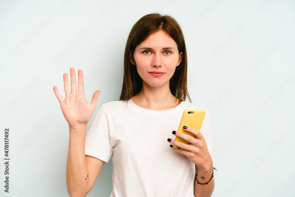Young English woman holding mobile phone isolated on blue background smiling cheerful showing number five with fingers. Young English woman holding mobile phone isolated on blue background smiling cheerful showing number five with fingers.