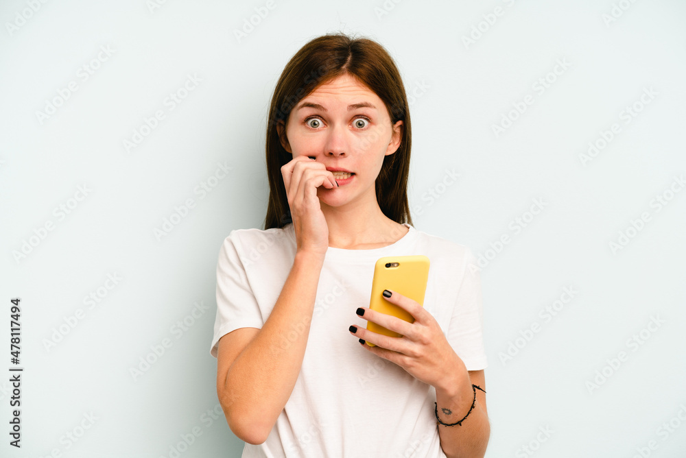 Young English woman holding mobile phone isolated on blue background biting fingernails, nervous and very anxious. Young English woman holding mobile phone isolated on blue background biting fingernails, nervous and very anxious.