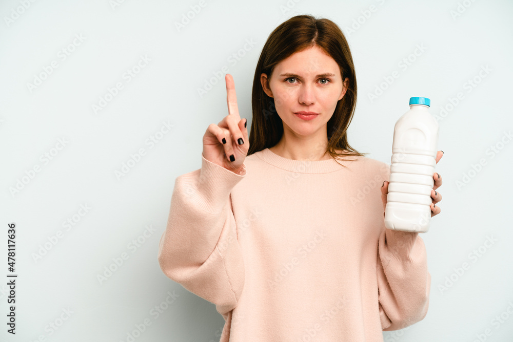 Young English woman holding a bottle of milk isolated on blue background showing number one with finger. Young English woman holding a bottle of milk isolated on blue background showing number one with finger.