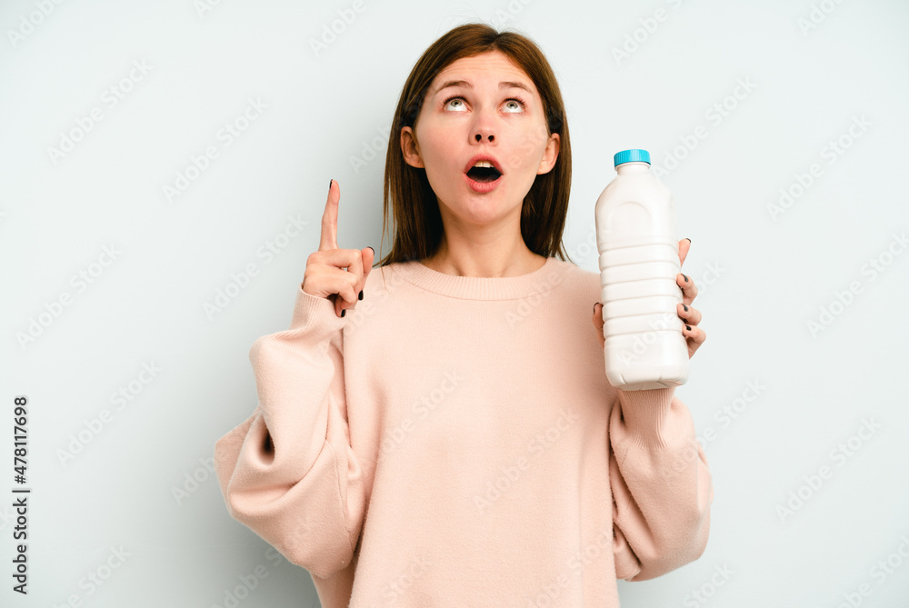 Young English woman holding a bottle of milk isolated on blue background pointing upside with opened mouth. Young English woman holding a bottle of milk isolated on blue background pointing upside with opened mouth.