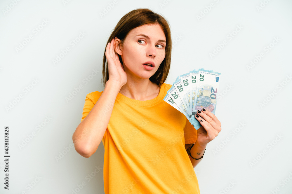Young English woman holding banknotes isolated on blue background trying to listening a gossip. Young English woman holding banknotes isolated on blue background trying to listening a gossip.