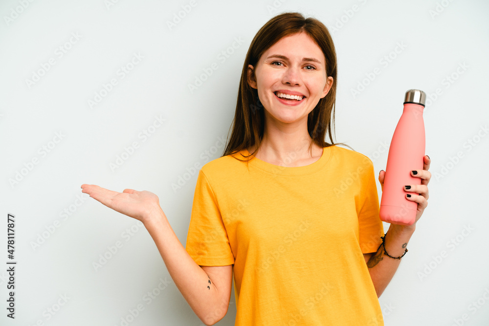 Young English woman holding a thermo isolated on blue background showing a copy space on a palm and holding another hand on waist. Young English woman holding a thermo isolated on blue background showing a copy space on a palm and holding another hand on waist.