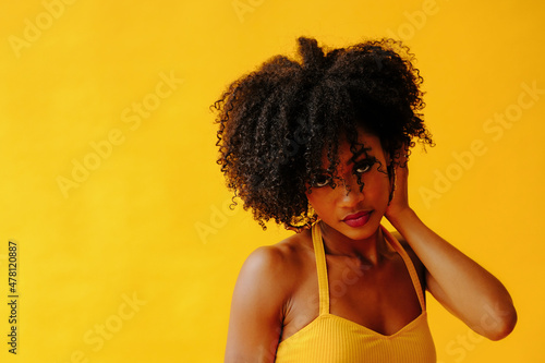 beautiful young African american woman with curly hair in bright summer outfit posing isolated on yellow background