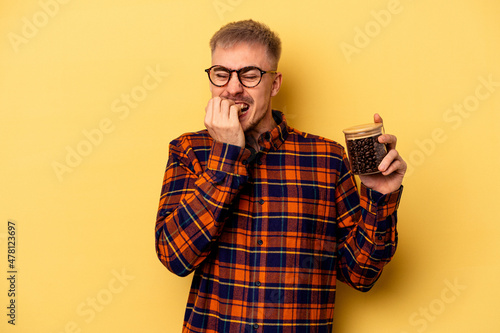 Young caucasian man holding coffee jar isolated on yellow background biting fingernails, nervous and very anxious. © Asier