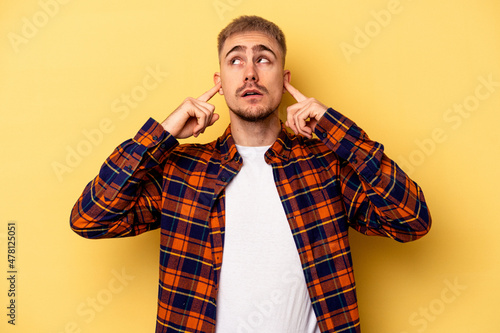 Young caucasian man isolated on yellow background covering ears with fingers, stressed and desperate by a loudly ambient. © Asier