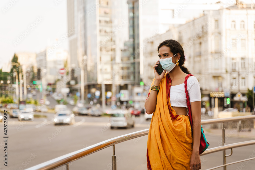 Obraz premium Young indian woman in face mask talking on cellphone at city street