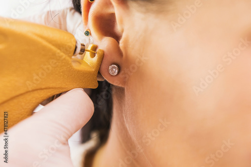 A doctor in sterile white medical gloves pierces the ears of a young girl in the medical office with a piercing gun. photo