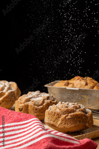 Close-up of individual plum cakes with falling sugar, with red kitchen towel, selective focus, black background, vertical