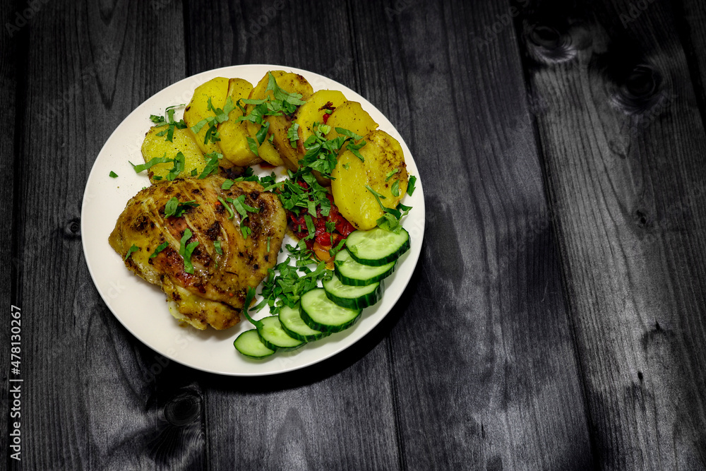 A dish with baked chicken and potatoes, garnished with green parsley and sliced cucumber, top view. On a dark wooden background. A dish with baked chicken and potatoes, garnished with green parsley and sliced cucumber, top view. On a dark wooden background.