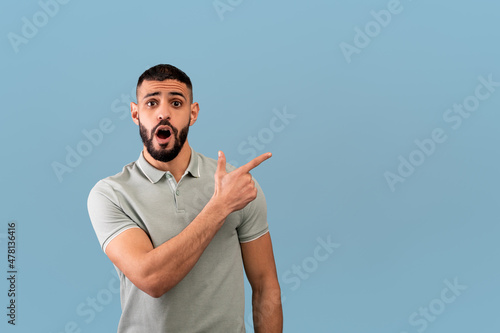 Surprised arab guy pointing aside at blank space and looking at camera with open mouth, posing over blue background photo