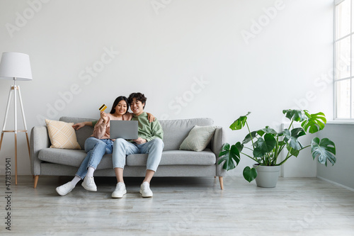 Fotografie, Obraz Asian couple using credit card and laptop, sitting on couch, ordering goods in w