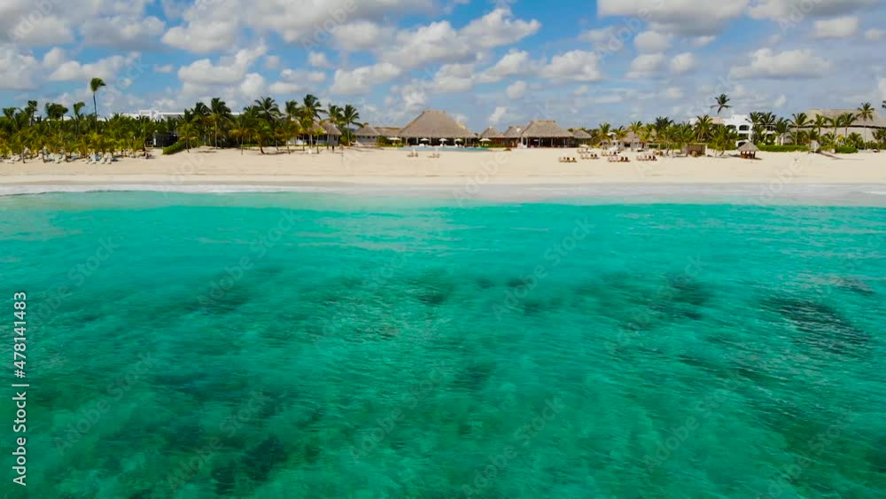Drone flying forward ander azure clear sea water. Aerial Moving Forward To Thatched Rooftop Building Surrounded By Palm Trees And Resort Buildings On Caribbean Beach. Aerial view of resort Punta Cana Drone flying forward ander azure clear sea water. Aerial Moving Forward To Thatched Rooftop Building Surrounded By Palm Trees And Resort Buildings On Caribbean Beach. Aerial view of resort Punta Cana
