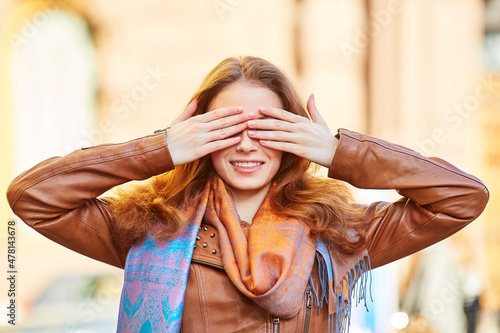 Young beautiful girl smiling and playing closes her eyes with her hands. Red-haired girl in a brown leather jacket on the street