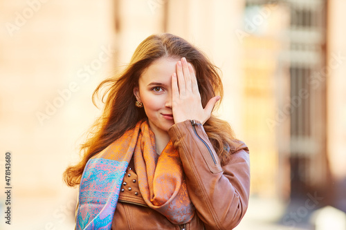 Young beautiful girl smiling and playing closes one eye with her hand. Red-haired girl in a brown leather jacket on the street
