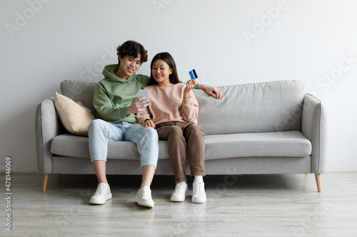 Cheery Asian couple with credit card and smartphone sitting on sofa, ordering goods on internet at home, copy space