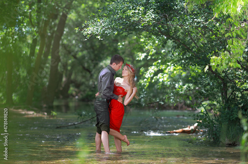 Fototapeta Naklejka Na Ścianę i Meble -  A guy and a beautiful young blonde girl with long hair in a red dress are having fun holding hands in the river water.