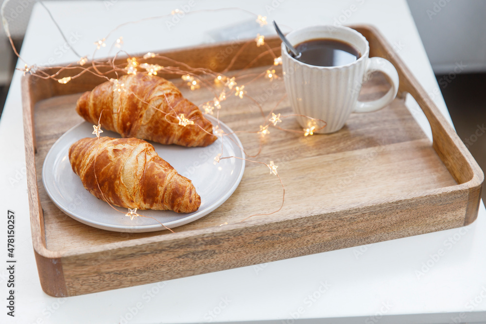Freshly baked croissant on a gray round plate, white cup with coffee and garland on a tray on the table Freshly baked croissant on a gray round plate, white cup with coffee and garland on a tray on the table