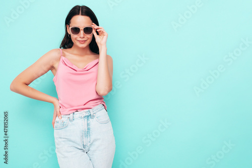 Portrait of young beautiful smiling female in trendy summer clothes. Sexy carefree woman posing near blue wall in studio. Positive model having fun indoors. Cheerful and happy. In sunglasses