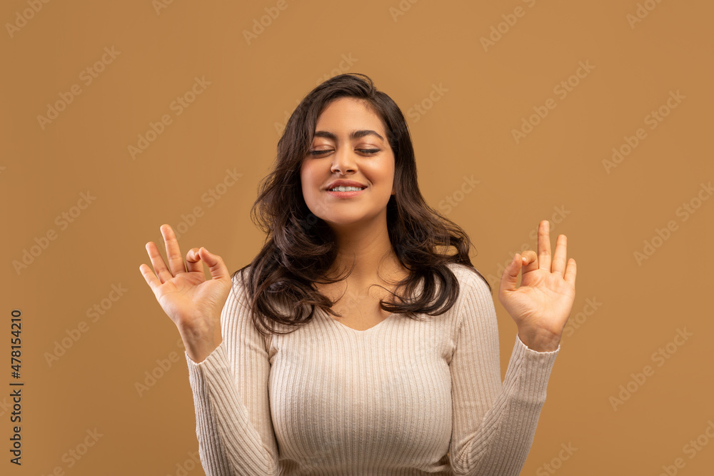 Meditation concept. Peaceful arab woman making om gesture and meditating with eyes closed, beige background - obrazy, fototapety, plakaty 