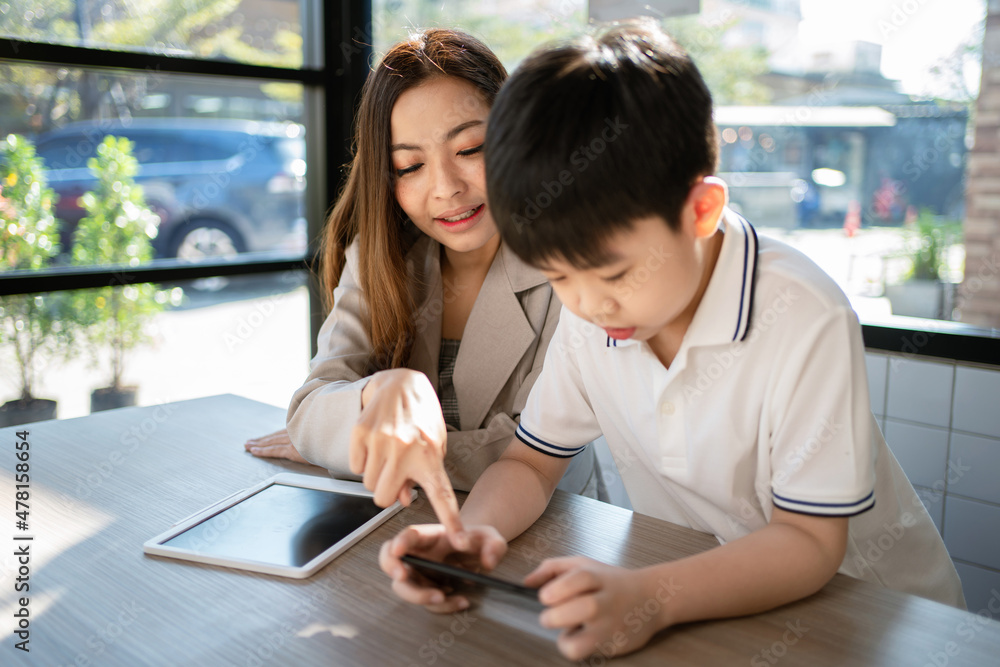 Woman point to the phone and talk with child. A boy playing game on the smartphone and sitting with his sister in the coffee shop or cafe. Learning from internet. Education and relaxation concept. Woman point to the phone and talk with child. A boy playing game on the smartphone and sitting with his sister in the coffee shop or cafe. Learning from internet. Education and relaxation concept.