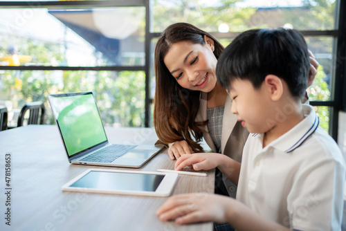 Businesswoman sitting beside kid and teach homework in the coffee shop. Woman stroking child's head and smile with copy space. Boy feeling happy during learning. Education and weekend concept.
