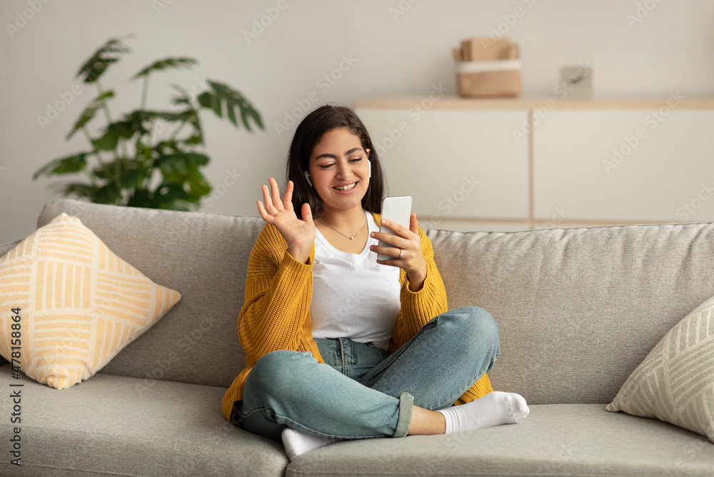 Happy arab lady video calling via cellphone, wearing headphones and waving hand while sitting on sofa at home Happy arab lady video calling via cellphone, wearing headphones and waving hand while sitting on sofa at home
