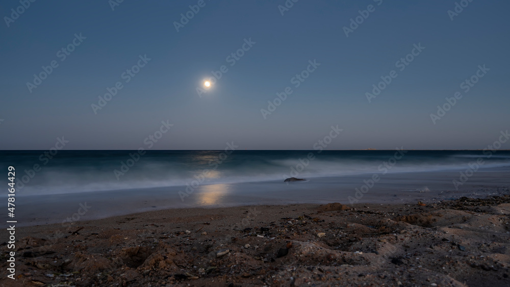A night on an Egyptian beach. The full moon is shining in the dark blue sky. Waves are foaming on the sand. Moonlight path on the water. Long exposure