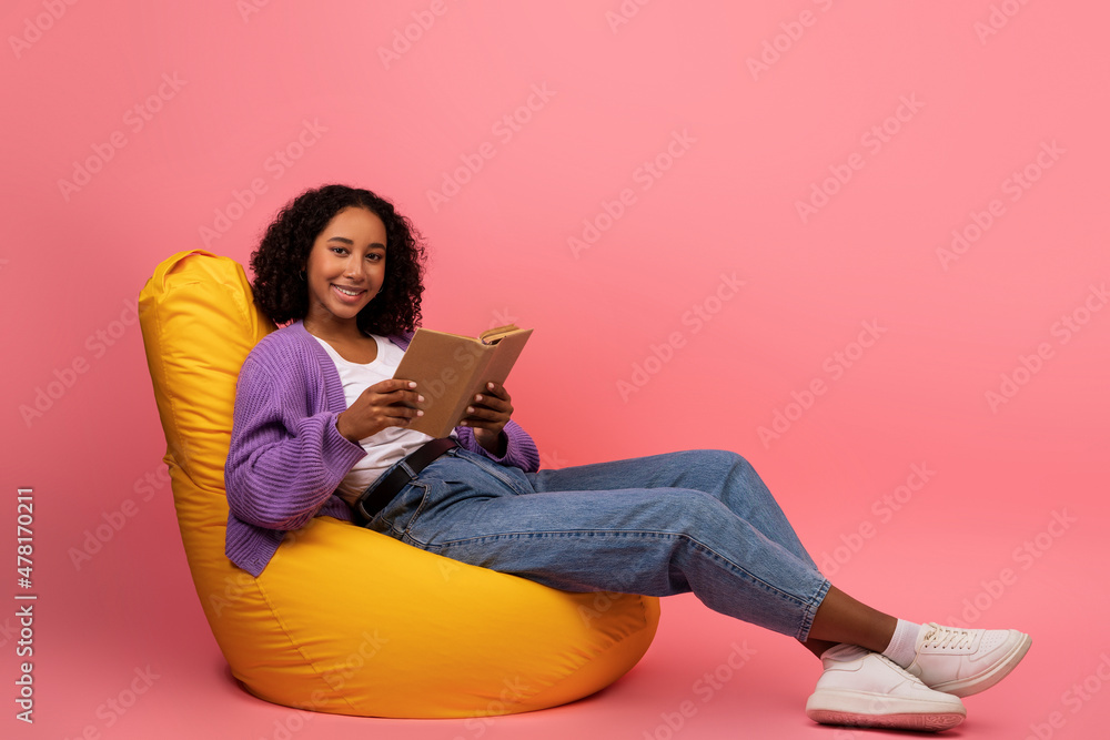 Weekend hobbies. Cheerful young black woman with open book sitting in comfy bean bag chair on pink background Weekend hobbies. Cheerful young black woman with open book sitting in comfy bean bag chair on pink background