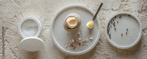 Canvas One boiled egg in stoneware egg cup with egg shell broken pieces in concrete tray, salt flakes and pepper in white concrete bowls on rough textured clay background