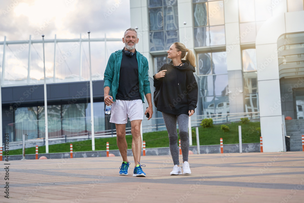 Full length shot of sportive middle aged couple in sportswear looking cheerful while walking together outdoors after workout