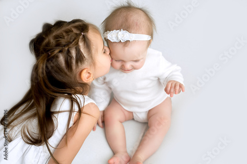 A cute three-year-old girl with pigtails kisses little sister gently on the cheek. Upbringing and mutual understanding in the family. Idyll in the relationship between children