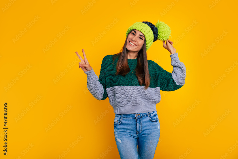 Happy cute young modern woman in hat shows victory sign while she posing isolated on yellow background and smiling Happy cute young modern woman in hat shows victory sign while she posing isolated on yellow background and smiling