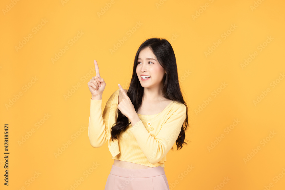 Beautiful Asian woman wearing yellow t-shirt smile and pointing finger at copy space isolated on yellow background studio portrait. Beautiful Asian woman wearing yellow t-shirt smile and pointing finger at copy space isolated on yellow background studio portrait.