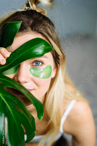 Vertical portrait of blond woman looking through monstera leaf with cute hairstyle and fresh soft skin and green patches looking at camera in bedroom interior.