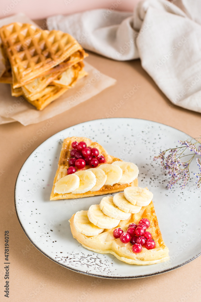Heart shaped Belgian waffles with red berries and banana on a plate on the table. Valentine's Day. Close-up. Vertical view Heart shaped Belgian waffles with red berries and banana on a plate on the table. Valentine's Day. Close-up. Vertical view