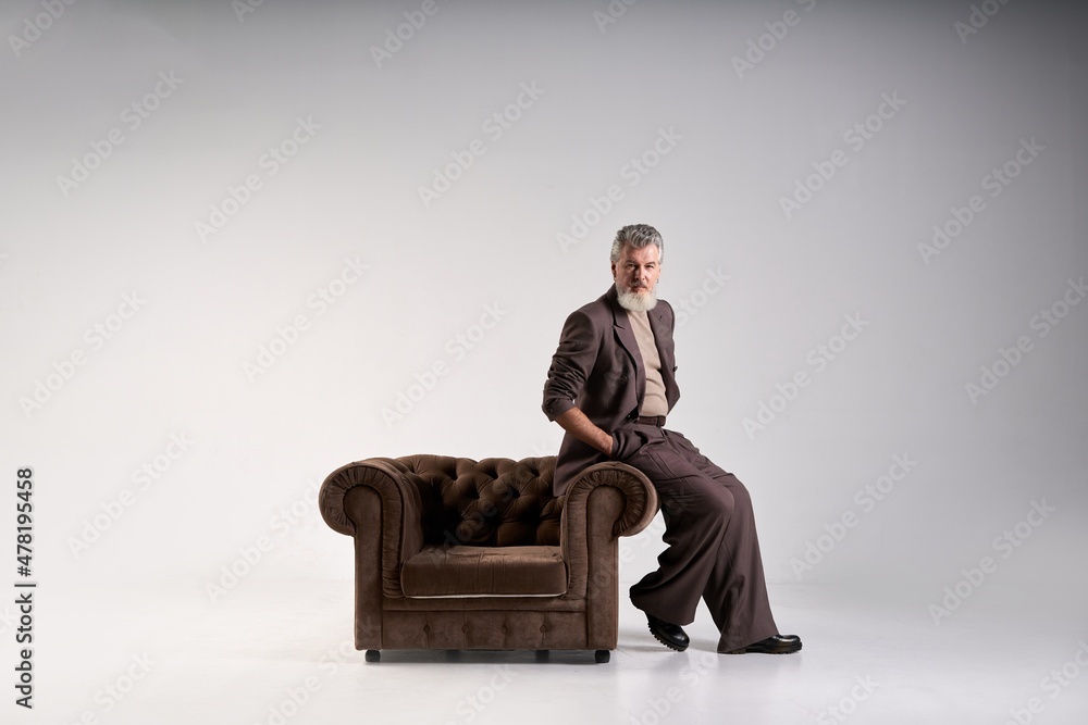 Full length shot of fashionable mature man with beard wearing elegant suit looking at camera, leaning on armchair while posing isolated over light background Full length shot of fashionable mature man with beard wearing elegant suit looking at camera, leaning on armchair while posing isolated over light background